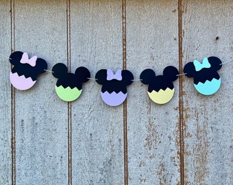 May include: A garland of five black and white Mickey Mouse heads with pastel colored accents. The heads are strung together with white string. The colors of the accents are light pink, light green, lavender, yellow, and light blue.