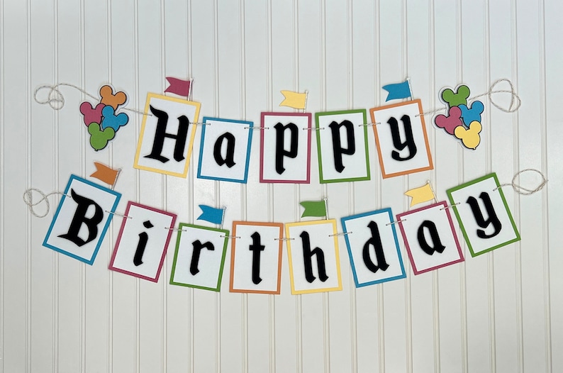 May include: A colorful Happy Birthday banner with Mickey Mouse ears and flags. The banner is made of paper and has a string for hanging.