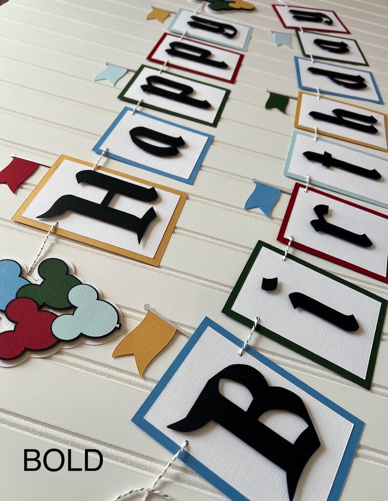 May include: A colorful banner with the words "Happy Birthday" in black letters on white squares with different colored borders. The banner is strung with white twine and has small flags with different colors.