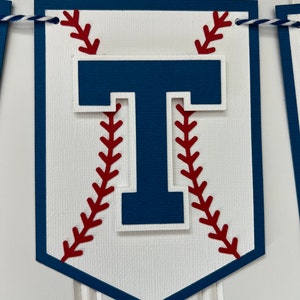 Pode incluir: Uma faixa branca com tema de beisebol com uma letra "T" azul no centro. A letra &eacute; cercada por costuras de beisebol vermelhas.