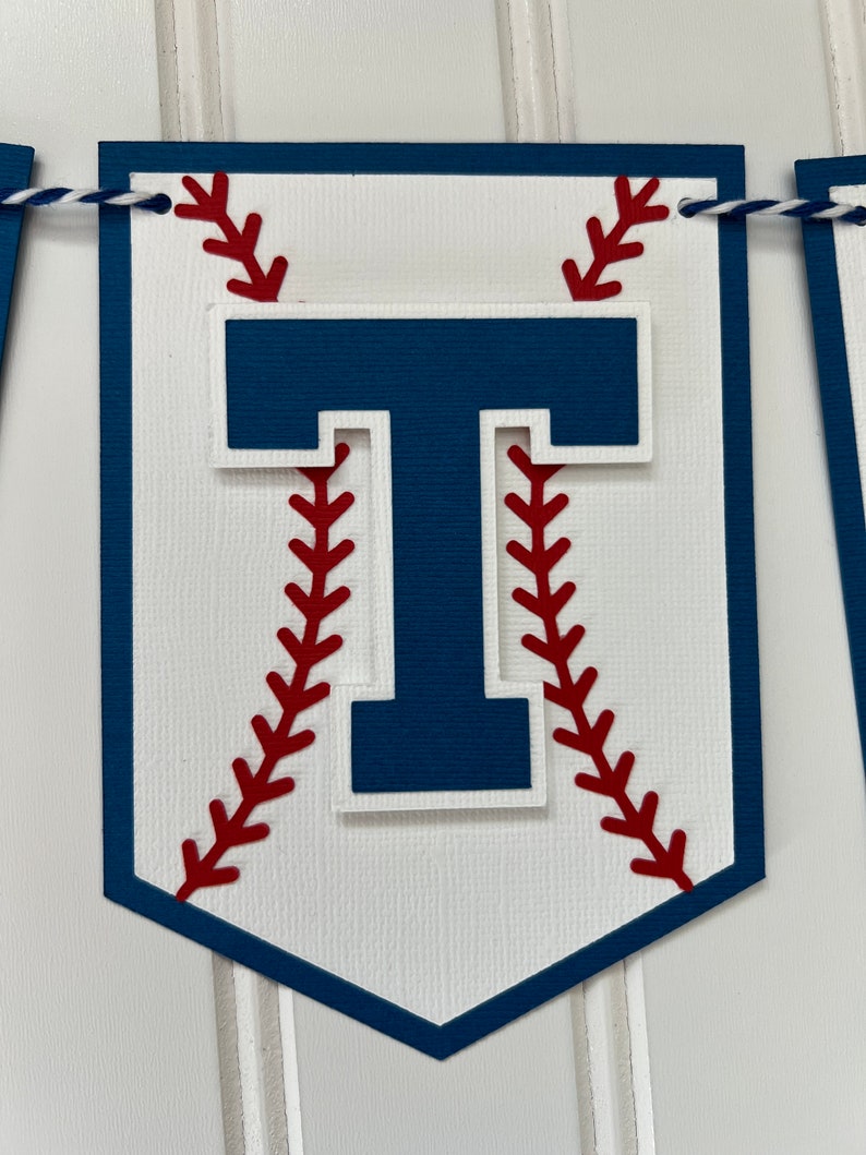 Pode incluir: Uma faixa branca com tema de beisebol com uma letra "T" azul no centro. A letra &eacute; cercada por costuras de beisebol vermelhas.
