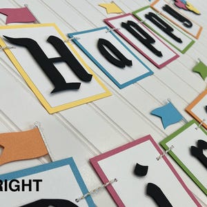 May include: A colorful Happy Birthday banner with black letters on white paper. The banner is made of different colored cardstock and has a string running through it. The letters are in a bold, gothic font.