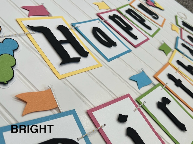 May include: A colorful Happy Birthday banner with black letters on white paper. The banner is made of different colored cardstock and has a string running through it. The letters are in a bold, gothic font.