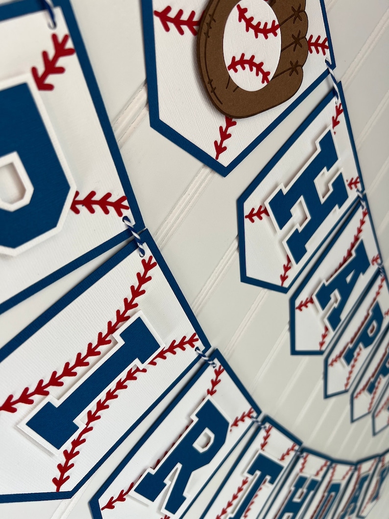 Pode incluir: Uma faixa com tema de beisebol com a palavra "Happy" em letras azuis sobre um fundo branco com costuras vermelhas. A faixa &eacute; decorada com uma luva de beisebol marrom e uma bola de beisebol branca com costuras vermelhas.