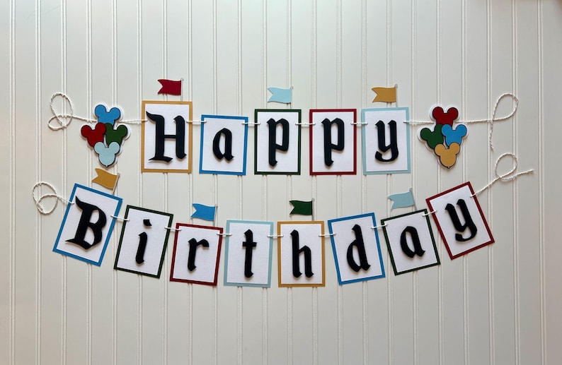 May include: A Happy Birthday banner with colorful flags and Mickey Mouse heads. The banner is made of white cardstock with black lettering and is strung on white twine.