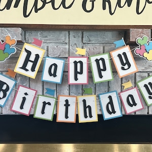 May include: A colorful banner with the words "Happy Birthday" in black letters on white squares. Each square has a different colored border. The banner is strung with a white string and has small flags hanging from it.