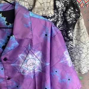 Può includere: Una camicia viola con bottoni, con un motivo geometrico e una chiusura con bottone nero. La camicia ha un colletto e maniche corte.