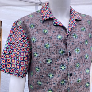 Puede incluir: Camisa de manga corta con un patrón geométrico único. La camisa presenta una base marrón con un diseño circular repetitivo y mangas y cuello contrastantes con un patrón de diamantes colorido. La camisa se exhibe en un maniquí blanco.
