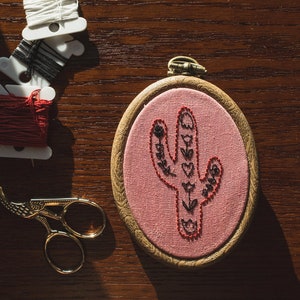 May include: An embroidery hoop with a pink fabric featuring a cactus design stitched in black and red thread. Beside it are spools of white, black, and red thread, along with gold scissors. The items are arranged on a wooden surface.
