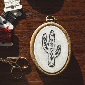 May include: An embroidered cactus design in a wooden hoop. The cactus is black and decorated with hearts and flowers. Gold scissors and spools of thread in white, black, and red are on a wooden surface.