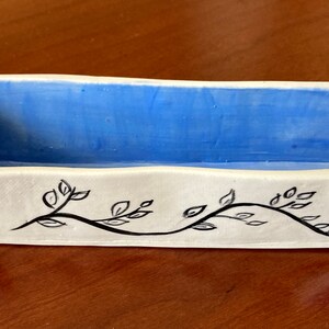 May include: A white ceramic rectangular dish with a blue glaze inside and a black floral design on the outside. The dish has a slightly raised rim.