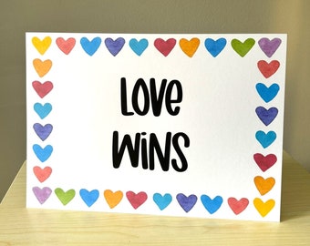May include: A white card with the words "LOVE WINS" in black lettering. The card is framed with a border of colorful watercolor hearts in shades of red, orange, yellow, green, blue, and purple. The card is displayed on a light wooden surface.