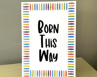 May include: A white rectangular print with the words "BORN THIS WAY" in black, framed by colorful vertical stripes. The print is displayed on a gold-colored metal stand, suitable for home decor.
