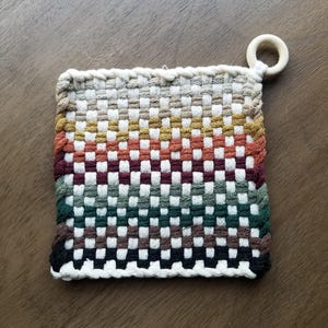 May include: A woven pot holder with a checkered pattern in shades of brown, red, green, and white. The pot holder has a wooden loop attached to the top corner.