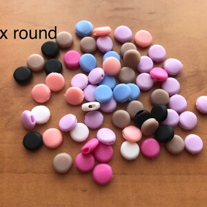 May include: A mix of round silicone beads in various colors, including black, brown, pink, purple, and blue. The beads are scattered on a wooden surface. Mix round.