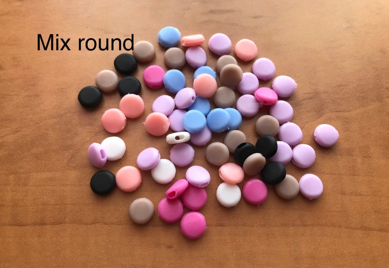 May include: A mix of round silicone beads in various colors, including black, brown, pink, purple, and blue. The beads are scattered on a wooden surface. Mix round.