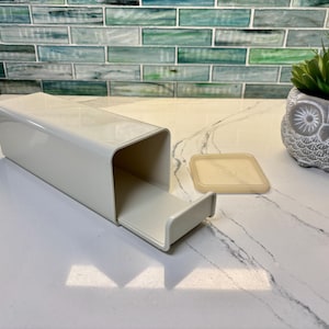 May include: A rectangular, off-white plastic storage container with a sliding tray and a separate, matching square lid. The container features a smooth, glossy finish and an open-ended design, providing a functional storage solution for small items.