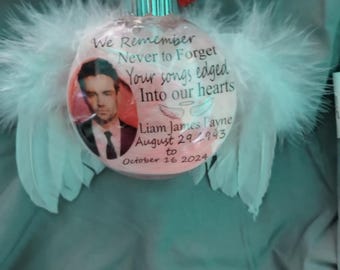May include: A clear glass Christmas bauble featuring a photograph of a man in a suit, adorned with white feathers. The bauble has the text "We Remember Never to Forget Your songs edged Into our hearts Liam James Payne August 29 1993 to October 16 2024."