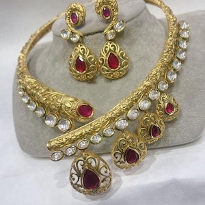 May include: A gold choker necklace with a decorative design and red and white stones. The necklace is paired with matching earrings with red and white stones.