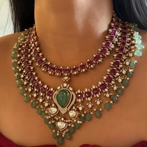 Puede incluir: Un collar de varias capas con piedras de color rubí, gemas verde esmeralda y cristales transparentes. El collar tiene un engaste dorado y un colgante con una gran piedra verde ovalada.