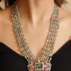 May include: A statement necklace and matching earrings. The necklace has multiple strands of green beads, red and white accents, and a large pendant with a central green stone. The pendant is surrounded by intricate metalwork.