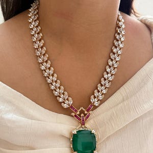 May include: A gold-toned necklace featuring a square emerald-green pendant. The necklace has a leaf-like design with clear, sparkling stones and red accents. The pendant is surrounded by smaller clear stones.