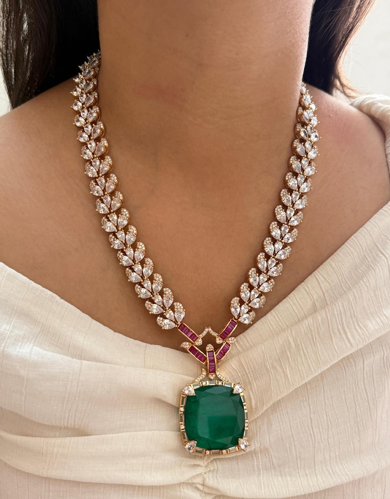 May include: A gold-toned necklace featuring a square emerald-green pendant. The necklace has a leaf-like design with clear, sparkling stones and red accents. The pendant is surrounded by smaller clear stones.