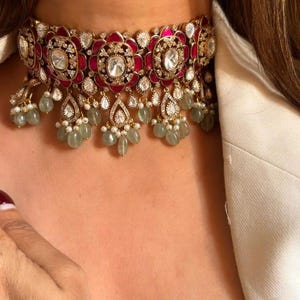 Op de afbeelding: Een gouden chokerketting met een rood en wit bloemenmotief en een groene edelsteen druppelhanger.