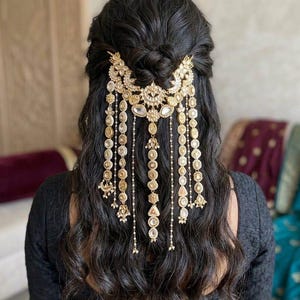 Könnte beinhalten: Ein goldfarbenes Haaraccessoire mit aufwendigen Details und herabhängenden Ketten. Das verzierte Stück weist ein zentrales Blumendesign und zahlreiche Anhänger mit klaren und goldenen Akzenten auf. Das Accessoire ist vor dunklem, welligem Haar platziert.