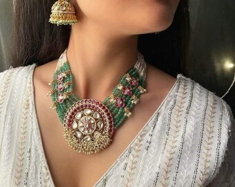May include: A gold and green beaded necklace with a large, round pendant featuring intricate floral designs and red and white stones. The necklace is worn by a person with a white blouse.