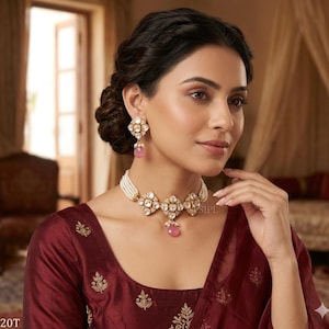 Könnte beinhalten: Eine Frau trägt eine goldfarbene Halskette und passende Ohrringe. Der Schmuck hat klare, quadratisch geschliffene Steine und rosa tropfenförmige Akzente. Die Halskette ist an einer Kette aus weißen Perlen befestigt. Die Frau trägt ein weinrotes Oberteil.