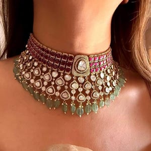 May include: A gold choker necklace with a central square pendant. The necklace features alternating sections of red and pink gemstones, with dangling green beads. The design includes numerous clear, round-cut stones.