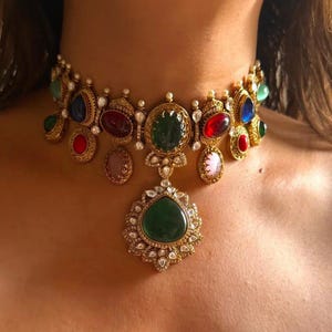 May include: A gold choker necklace with a variety of colourful gemstones. The necklace features a central pendant with a large green stone and smaller, teardrop-shaped pendants. The gemstones are in shades of red, green, blue, and pink.