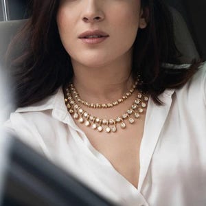 Puede incluir: Una mujer con una camisa blanca y un collar de oro y diamantes. El collar tiene dos hileras de peque&ntilde;os diamantes redondos y una hilera de diamantes redondos m&aacute;s grandes que cuelgan de la parte inferior.