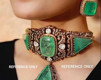 May include: A gold choker necklace with a large emerald green gemstone and smaller diamonds. The necklace has a pendant with a teardrop-shaped emerald green gemstone and diamonds. The necklace is on a model's neck. REFERENCE ONLY.
