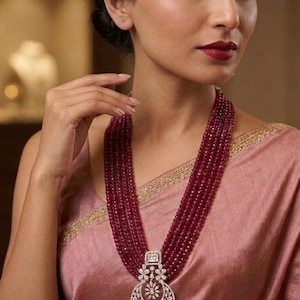 May include: A multi-strand ruby bead necklace with a large, ornate pendant. The pendant features a floral and geometric design with clear gemstones. The necklace is displayed on a person wearing a pink garment.