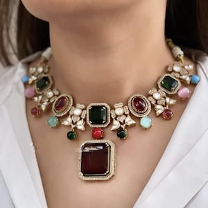 May include: A gold-toned necklace featuring a large, rectangular, dark red gemstone pendant. The necklace has a series of colorful gemstones, including green, red, pink, and blue, set in a floral design with clear crystals.