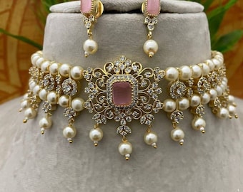 Può includere: Un girocollo in oro con un centro in pietra rosa e dettagli in perle. Il girocollo è abbinato a orecchini coordinati con pietre rosa e perle.