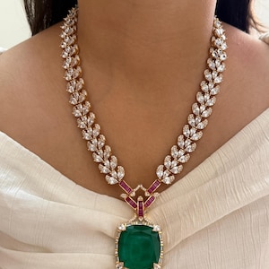 Peut inclure: Un collier doré avec un grand pendentif carré en émeraude verte. Le collier présente un motif de feuilles avec des pierres claires et scintillantes et des accents rouges. Le collier est porté autour du cou.
