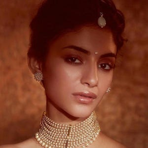 Könnte beinhalten: Eine Frau trägt eine goldene und perlmuttfarbene Halskette mit einem weißen Stein auf der Stirn. Sie hat dunkles Haar und dunkle Augen.