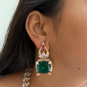 May include: Gold-toned earrings featuring a large, square, emerald-green gemstone at the center. The earrings are adorned with smaller, clear and red gemstones. The design includes a decorative top section with additional gemstones.