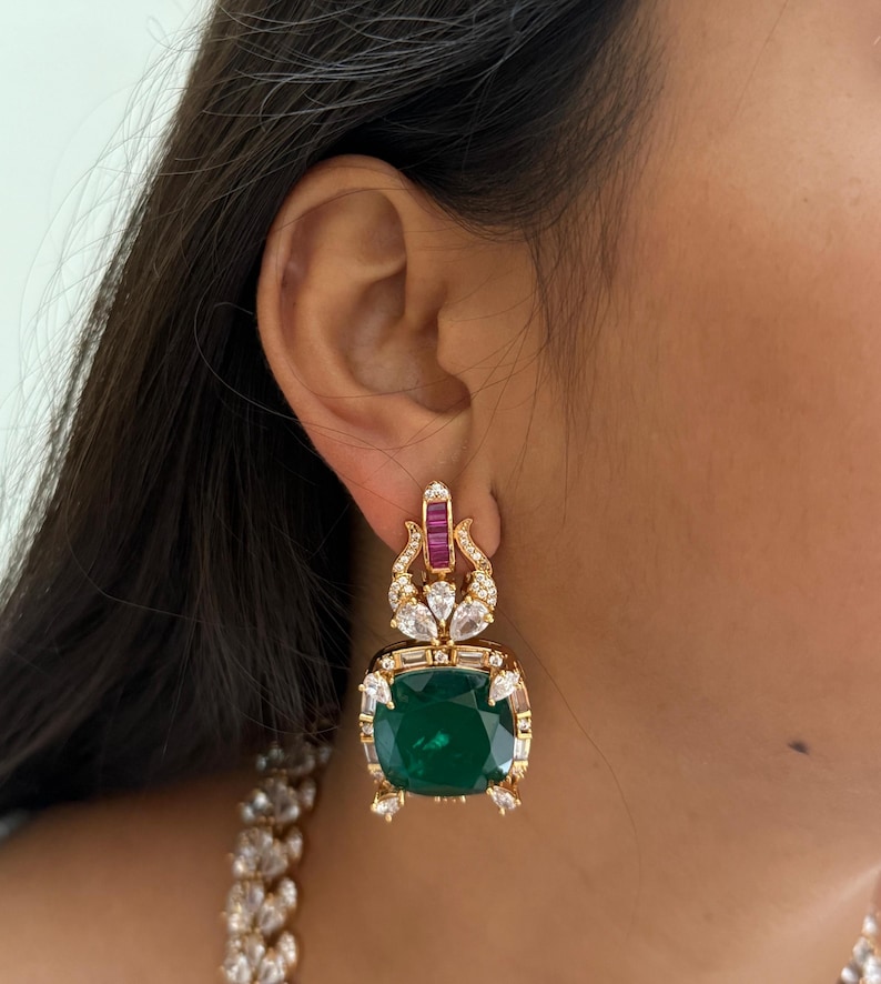 May include: Gold-toned earrings featuring a large, square, emerald-green gemstone at the center. The earrings are adorned with smaller, clear and red gemstones. The design includes a decorative top section with additional gemstones.
