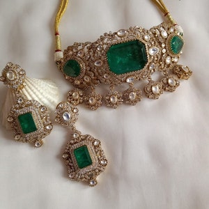 May include: A gold necklace and earrings set with emerald green gemstones and white stones. The necklace is a choker style with a delicate design and the earrings are long and dangle.