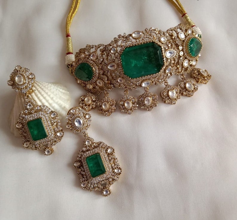 May include: A gold necklace and earrings set with emerald green gemstones and white stones. The necklace is a choker style with a delicate design and the earrings are long and dangle.