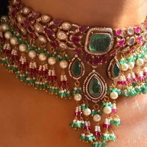 Puede incluir: Primer plano de un collar ornamentado con detalles de esmeraldas verdes, rubíes rojos y perlas. El collar tiene una base de metal dorado con detalles intrincados y múltiples capas de piedras preciosas y perlas, creando un aspecto lujoso.