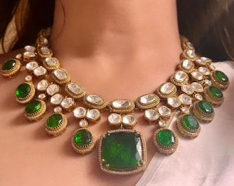 May include: A gold necklace with a large green gemstone pendant and smaller green gemstones. The necklace is made with white stones and has a layered design.