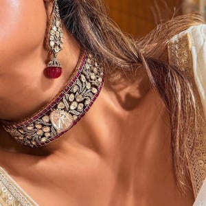 May include: An ornate necklace and matching earring set. The jewelry features a combination of clear and red gemstones set in a gold-colored metal. The necklace is a choker style, and the earring is a long drop style.