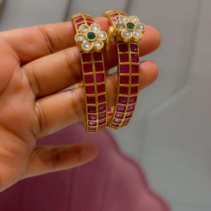 May include: Two gold bangles featuring a grid of square-cut red stones. Each bangle is accented with a floral-shaped centerpiece containing clear white stones surrounding a single round green stone in the center, set against a metallic gold frame.