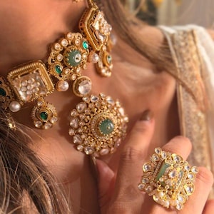 May include: Close-up of a gold-coloured jewellery set featuring a necklace, earrings, and rings. The pieces are adorned with clear and emerald-green gemstones and small pearls. The jewellery has an ornate, vintage design.