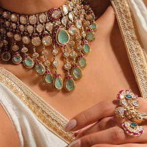 May include: A multi-layered gold choker necklace featuring teardrop-shaped light green stones, small round white stones, and accents of deep red stones. The set includes a matching ornate ring and a bracelet with square light green stones.